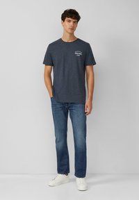 Marineblauwe t-shirt met ronde hals en korte mouwen, voorzien van een wit logo op de borst, gecombineerd met blauwe jeans en witte sneakers.