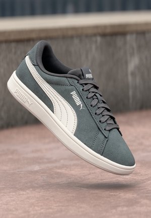 Szaro-białe trampki Puma z sznurówkami unoszące się nad różowawą betonową powierzchnią i szarą ścianą w tle.
