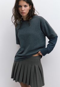 Kobieta z mokrymi włosami ubrana w warstwowe szare koszule i plisowaną szarą spódnicę, stojąca na jednolitym jasnym tle.