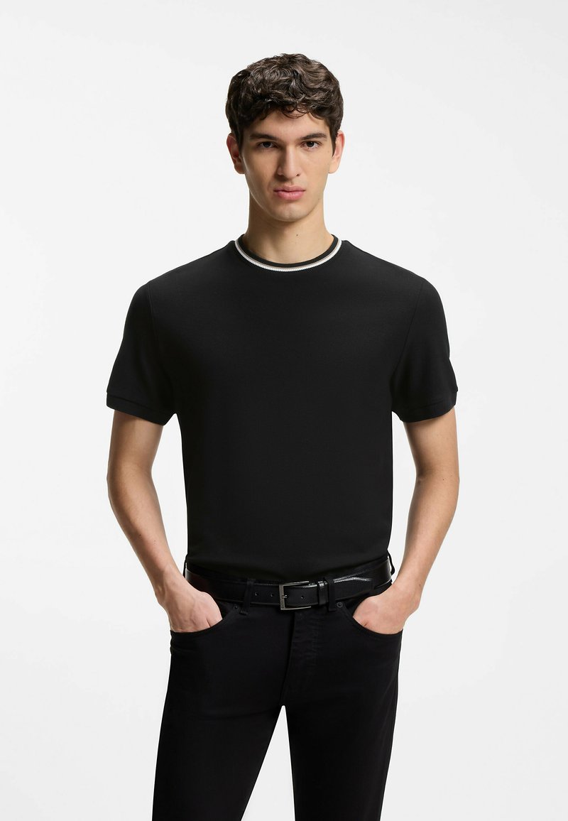 Jeune homme aux cheveux courts et bouclés portant un t-shirt noir avec un col bordé de blanc, un pantalon noir, les mains dans les poches, sur un fond blanc uni.