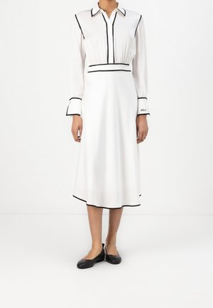Robe midi blanche à manches longues avec bordure noire, col pointu, boutonnage devant, détails aux poignets et chaussures plates noires sur une personne debout.