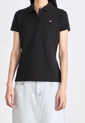 Vrouw die een zwart poloshirt met korte mouwen draagt met een klein rechthoekig logo op de borst en een lichte denim rok.