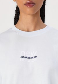 Balta kokvilnas T-krekls ar apaļu kakla izgriezumu, kuram uz krūtīm ir stilizēts "new balance" logo zilo un baltu drukas krāsā. Gluda tekstūra, brīvs piegriezums.