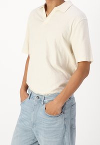 Camisa polo de manga corta de color crema con una superficie texturizada y un diseño de cuello en V, combinada con unos vaqueros de denim azul claro.