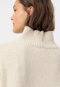 Beige gestrickter Pullover mit hohem Rippkragen und weicher Textur, sichtbaren Maschen und lockerer Passform auf der Rückseite.