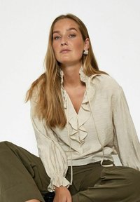 Light beige, textured blouse with ruffles along the neckline and cuffs; paired with olive green, loose-fitting trousers.