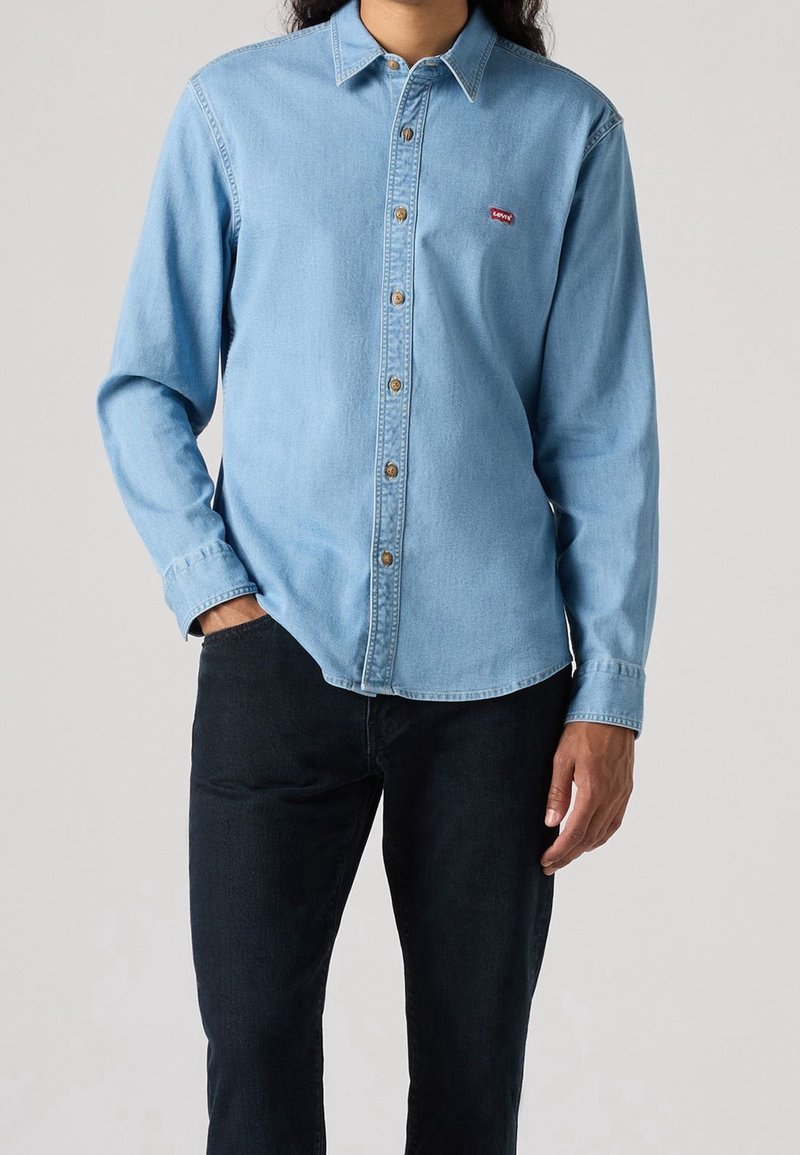 Personne portant une chemise en jean bleu clair à manches longues avec des boutons et un petit logo rouge Levi's sur la poitrine, associée à un pantalon noir.