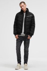 Mann mit Tätowierungen, der eine schwarze Camp David Steppjacke, einen hellgrauen Pullover, dunkle Jeans und weiße Turnschuhe trägt und vor einem weißen Hintergrund steht.