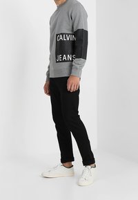 Šedá mikina s černými koženými rukávy, s bílým logem "CALVIN JEANS." V kombinaci s černými kalhotami a bílými teniskami.