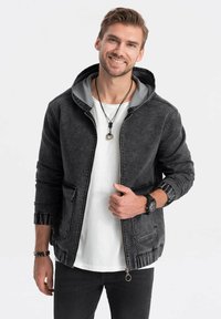 Giubbotto di jeans nero con cappuccio grigio, zip frontale, doppie tasche, polsini arricciati, texture vissuta, look sfruttato abbinato a una t-shirt bianca e jeans scuri.