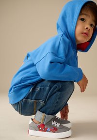 Sweat à capuche bleu avec doublure rouge, enfant accroupi en jean denim, baskets grises avec design Spider-Man, texture subtile dans le tissu.