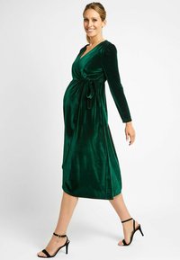 Wickeldress aus reichhaltigem smaragdgrünen Samt mit langen Ärmeln, gebundenem Taillengurt und knielangem Saum, kombiniert mit schwarzen Riemchen-Heels. Glatte Textur.