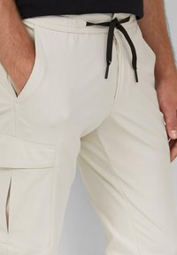 Beige cargobroek van lichtgewicht stof. Voorzien van een elastische taille met zwarte koorden, zijzakken en een splitdetail op het been.