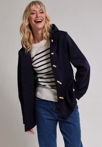 Mörkblå jacka med träknappar, med huva och avslappnad passform. Bärs över en krämfärgad skjorta med svarta ränder och blå jeans.