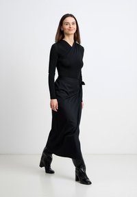 Kobieta pewnym krokiem, ubrana w czarną, długorękawową bluzkę kopertową, dopasowaną midi spódnicę oraz czarne botki na obcasie, na jednolitym białym tle.
