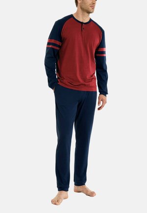 Uomo in piedi a piedi nudi, indossa una maglia raglan a maniche lunghe rossa e blu navy con righe sulle maniche e pantaloni blu navy, su sfondo bianco.