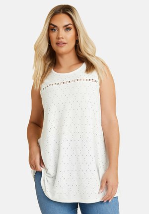 Blondynka w białej, bezrękawnikowej bluzce z małymi perforacjami i poziomym koronkowym detalem, w połączeniu z niebieskimi dżinsami.