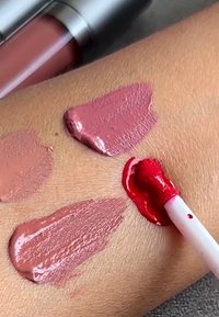 Leppeglans-applikator som påfører en sterk rød fargeprøve på huden, med tre andre beige og rosa leppefargeprøver i nærheten.