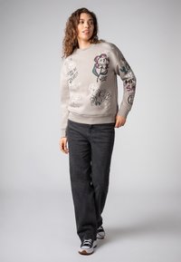 Woman with curly hair wearing a beige sweatshirt with colorful doodles and black jeans, standing against a plain light gray background.