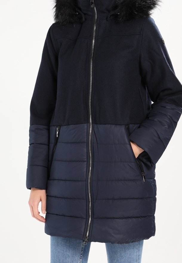Manteau bleu marine avec fermeture éclair, capuche en fausse fourrure, mélange de textures lisses et matelassées, avec poches latérales.