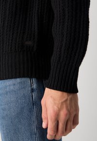 Close-up of a hand by the side wearing blue jeans and a black ribbed sweater with a small "BOSS" logo patch.