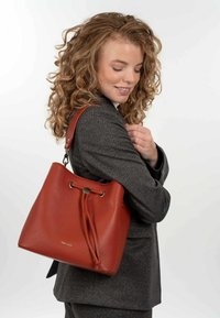 Borsa a secchiello rossa in pelle con superficie testurizzata, chiusura a coulisse e una tracolla beige, presentata su uno sfondo bianco.