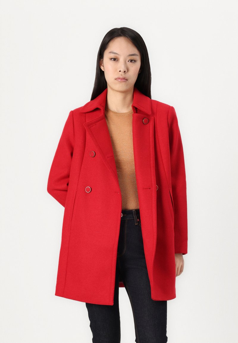 Roter doppelreihiger Mantel mit Reverskragen, großen Knöpfen und Taschen. Über einem braunen Pullover und dunklen Jeans getragen, die eine gerade Schnittführung haben.