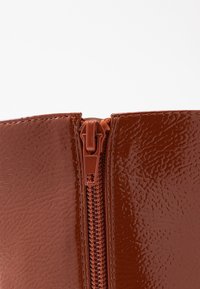 Bottines en cuir verni marron avec une surface lisse et brillante, dotées d'une fermeture éclair et d'un design structuré sur le haut.