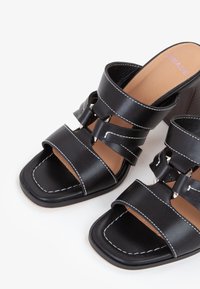 Sorte højhælede sandaler med to brede stropper, der har dekorerede udsparinger og hvid syning, kvadratisk tådesign og en brun indersål.