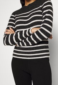 Personne portant un pull rayé horizontalement noir et blanc, les bras croisés sur une jupe ou un pantalon noir côtelé.