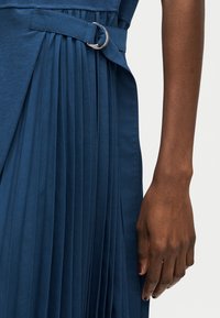 Σκούρο δέρμα χεριού δίπλα σε πλισέ φούστα ναυτικού μπλε χρώματος με ζώνη που έχει ασημένια αγκράφα διπλού δακτυλίου.