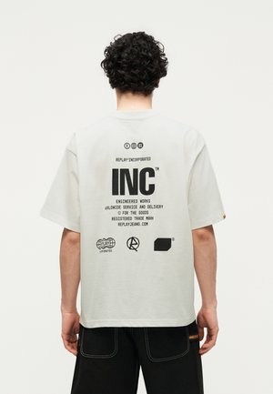 Hombre con cabello rizado que lleva una camiseta blanca oversize con texto y logos negros en la espalda, combinada con pantalones negros con costuras blancas.