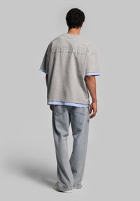 Gray oversized T-shirt with “WRSTBHVR” embossed on the back, featuring blue striped cuffs. Paired with light blue denim jeans.