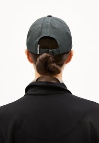 Persoon met donker haar in een lage knot, een donkergrijze baseballpet en een zwart jack, gezien van achteren tegen een effen achtergrond.