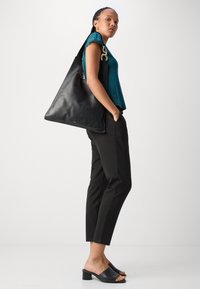 Sac tote en cuir noir avec une texture lisse et un accent en chaîne dorée proéminent. Le modèle porte un haut en teal, un pantalon noir et des mules à talons noirs.