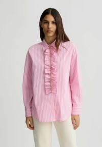 Chemise rayée rose et blanche à manches longues, avec un col boutonnière et un détail central en volants orné d'accents métalliques.