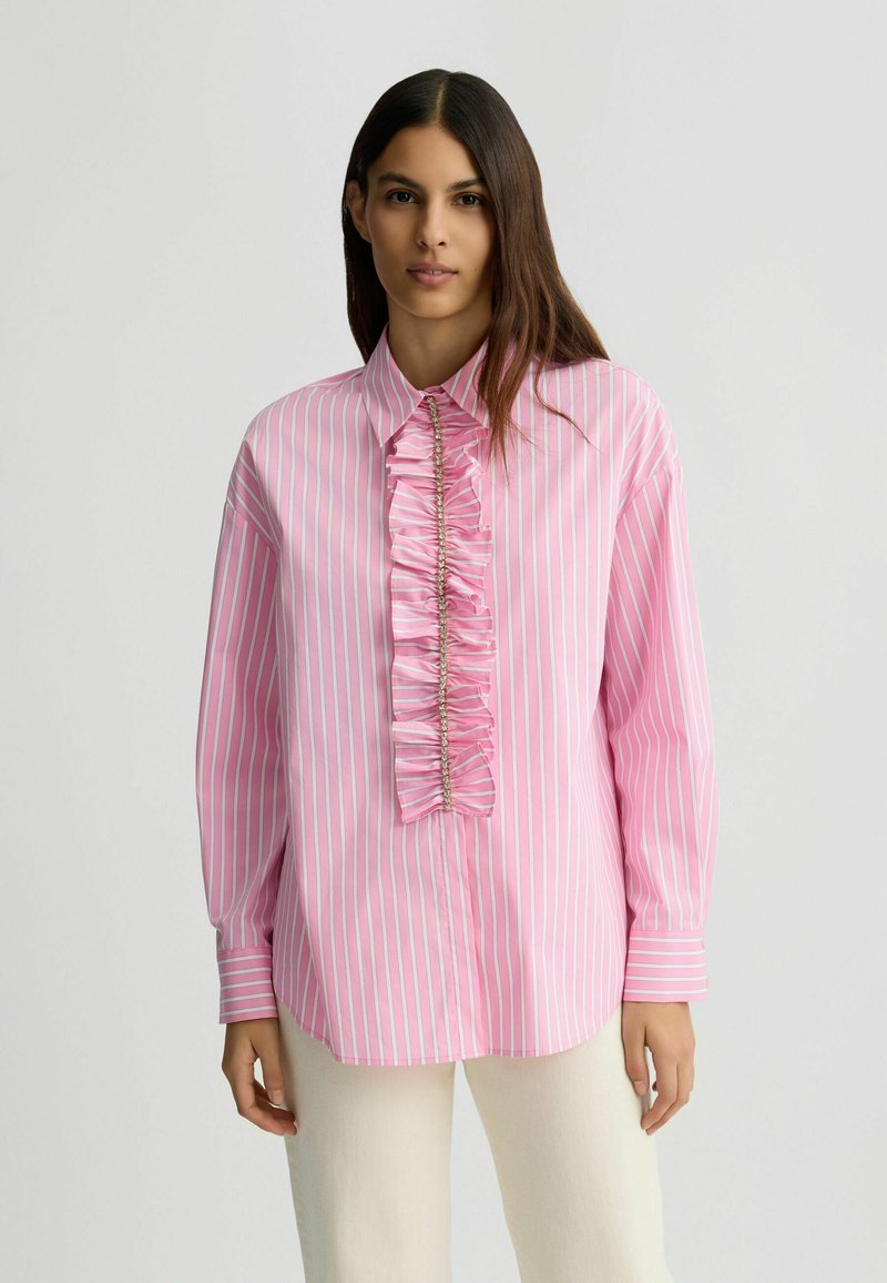 Chemise rayée rose et blanche à manches longues, avec un col boutonnière et un détail central en volants orné d'accents métalliques.