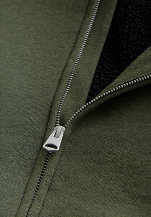 Grüne Zip-Jacke mit silbernem Reißverschluss, ausgestattet mit weichem schwarzen Fleece-Futter und strukturiertem Stoff. Nahaufnahme zur Hervorhebung der Details.