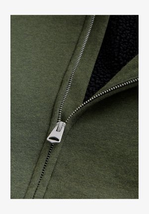 Grüne Zip-Jacke mit silbernem Reißverschluss, ausgestattet mit weichem schwarzen Fleece-Futter und strukturiertem Stoff. Nahaufnahme zur Hervorhebung der Details.