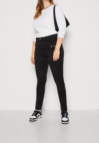 Jeans ajustados negros con textura acanalada en los muslos y bolsillos con cremallera, combinados con una camiseta blanca de manga larga acanalada y zapatillas negras.