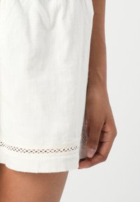 White shorts made of textured fabric, featuring a hem with a delicate lace trim. The shorts are fitted at the waist.