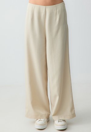Pantalon beige à jambes larges porté avec des baskets blanches, montré de la taille jusqu'aux pieds sur un fond clair uni.