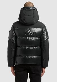 Chaqueta acolchada negra con acabado brillante, con capucha, diseño acolchado y costuras visibles. Combinada con pantalones negros, vista desde atrás.