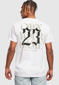 T-shirt blanc en coton avec un texte noir et gris audacieux au dos : "PROPRIÉTAIRE DE VOTRE JEU", "23" et "FAITES-LE LÉGENDAIRE". Manches courtes, coupe décontractée.