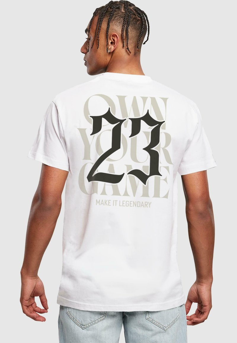 T-shirt blanc en coton avec un texte noir et gris audacieux au dos : "PROPRIÉTAIRE DE VOTRE JEU", "23" et "FAITES-LE LÉGENDAIRE". Manches courtes, coupe décontractée.