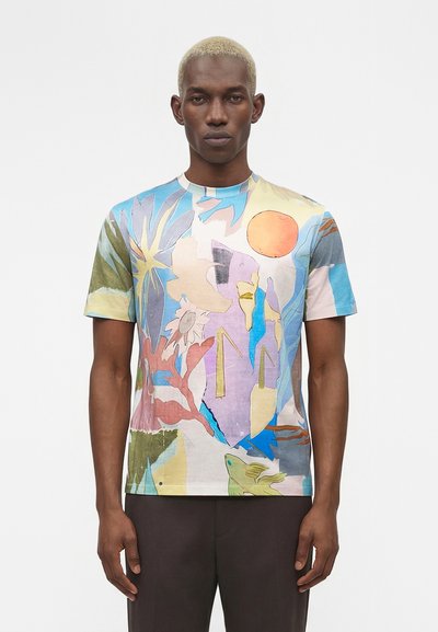 Homme aux cheveux blonds courts portant un t-shirt à manches courtes avec un imprimé abstrait multicolore et un pantalon foncé, debout devant un fond uni.