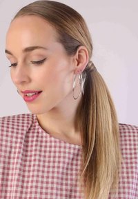 Model met lang haar in een strakke staart, draagt grote zilveren oorbellen. Ze draagt een roze geblokte top met een ronde hals.