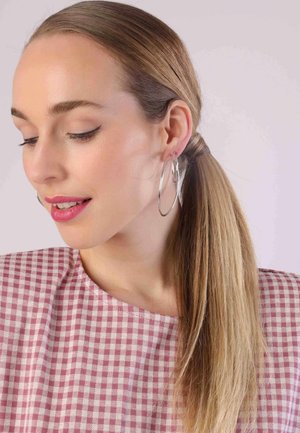 Model met lang haar in een strakke staart, draagt grote zilveren oorbellen. Ze draagt een roze geblokte top met een ronde hals.