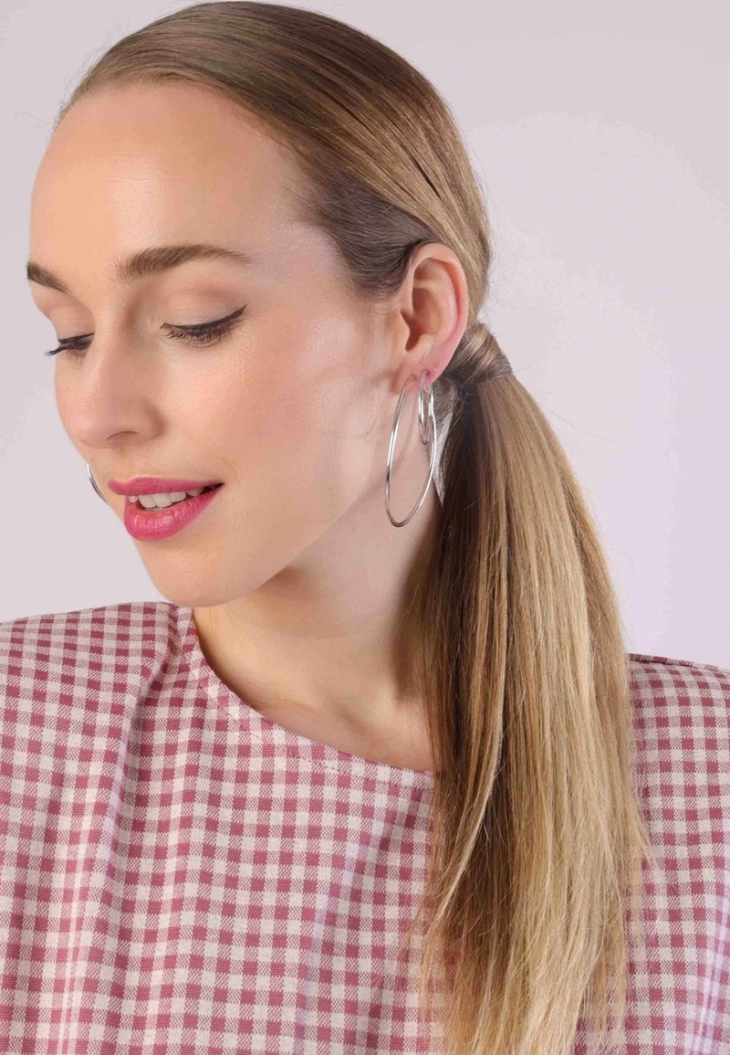 Model met lang haar in een strakke staart, draagt grote zilveren oorbellen. Ze draagt een roze geblokte top met een ronde hals.