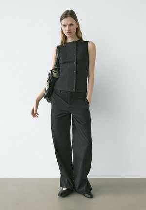 Žena stojící u bílé zdi, oblečená v černém bezrukávovém topu zapínaném na knoflíky, širokých černých kalhotách, černých botách a nesoucí černou tašku s šálou.